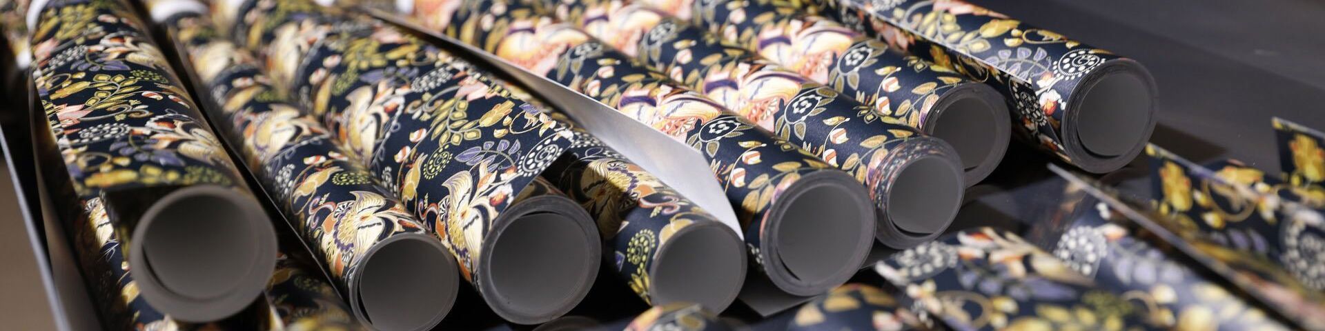 Eight rolls of wallpaper (and more just out of shot), photographed side by side, from the end of the rolls. They are patterned with leaves and flowers on a black background.